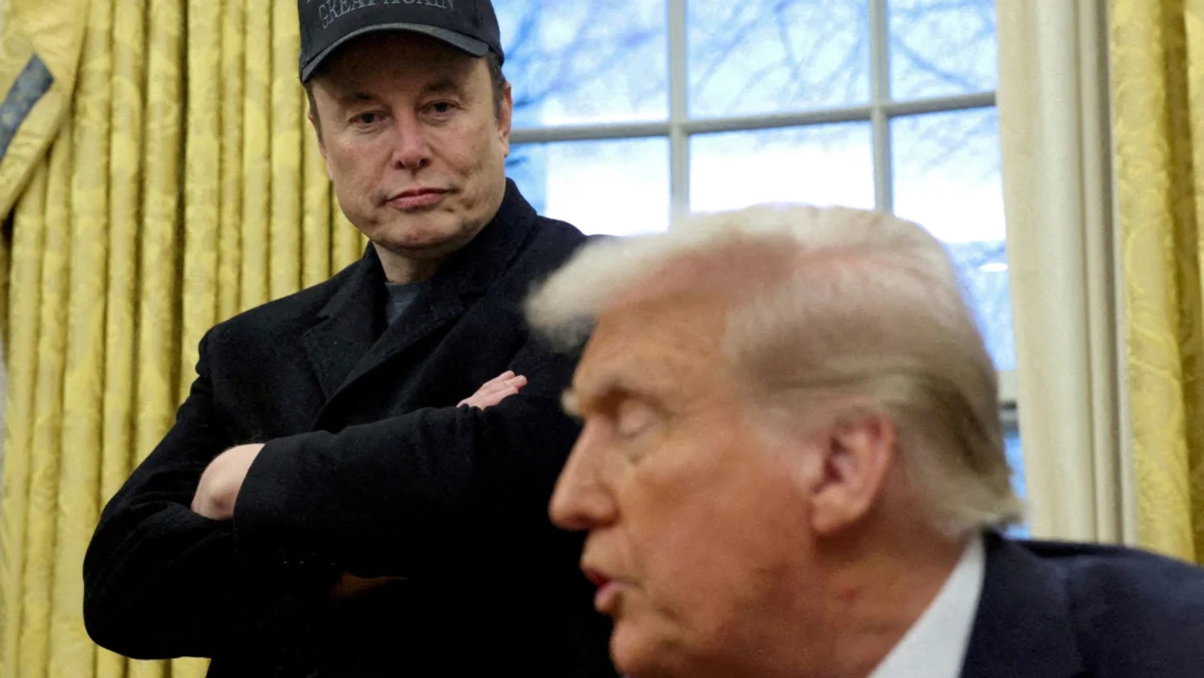 Elon Musk watches Donald Trump speak during a White House meeting, wearing a cryptic expression and a hat that reads “Great Again”