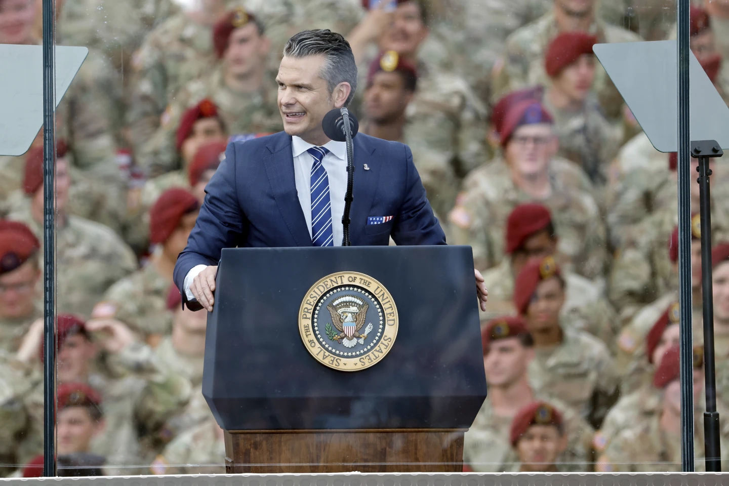 Pete Hegseth speaks at podium with the presidential seal, surrounded by troops in camouflage uniforms, during a military event supporting Trump’s base renaming plan