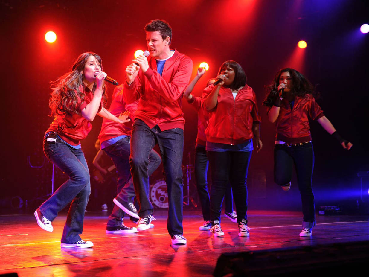 Cast members in red jackets perform on stage under dramatic lighting during a musical number
