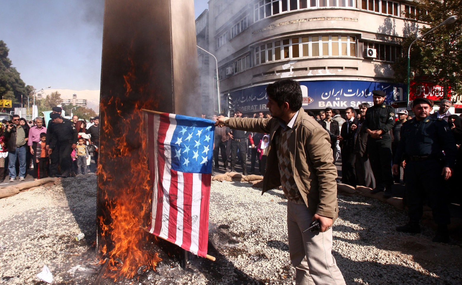 A Middle Eastern merchant surrounded by people in the street as he burns American flags labeled “flammable”