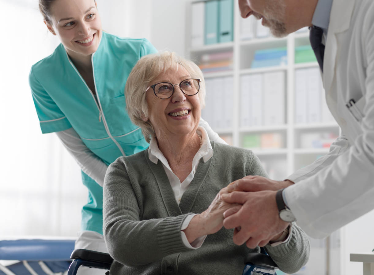 Smiling senior woman in wheelchair shakes hands with a doctor during a checkup, with a nurse standing behind her