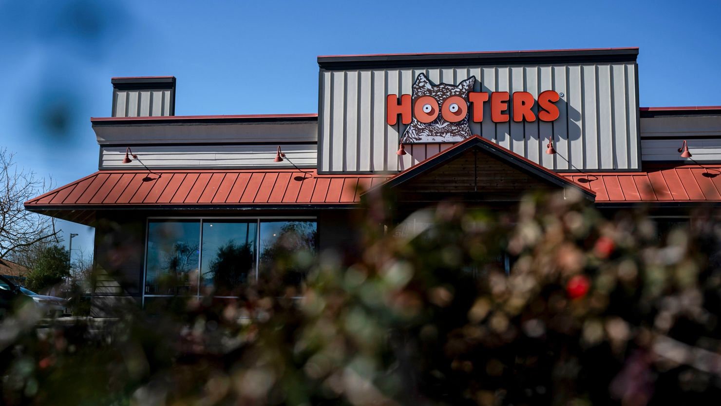 Hooters restaurant with its iconic orange sign seen from behind some bushes, as the chain closes 30 locations nationwide due to shifting family dining preferences.