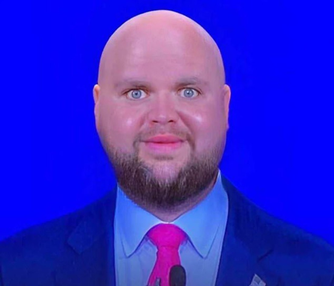 Close-up of a bald man with a trimmed beard and a slightly unsettling smile in front of a bright blue background