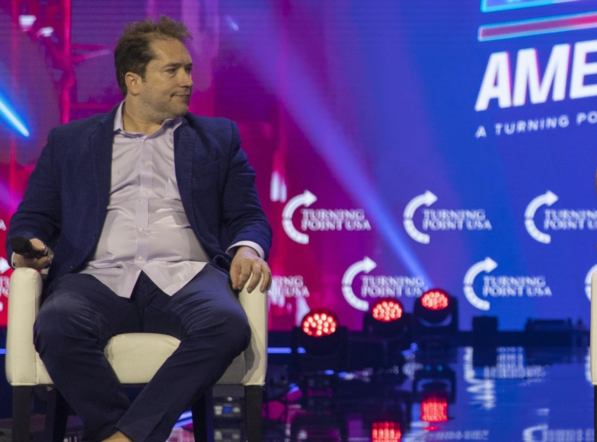 Man in blue suit holding microphone at Turning Point USA event, sitting on stage with colorful background