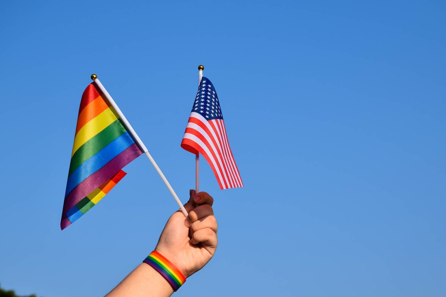 Hand holding a rainbow Pride flag and a U.S. flag against a blue sky
