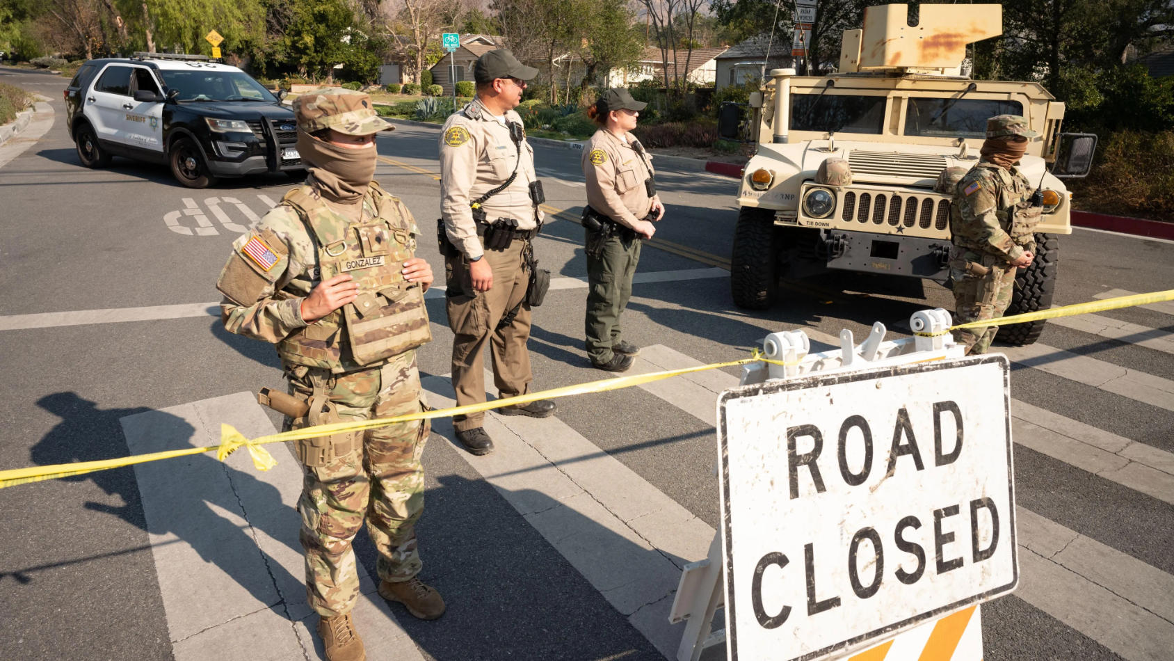 National Guard and sheriff deputies stand at a roadblock with a “Road Closed” sign and military vehicles in a suburban neighborhood