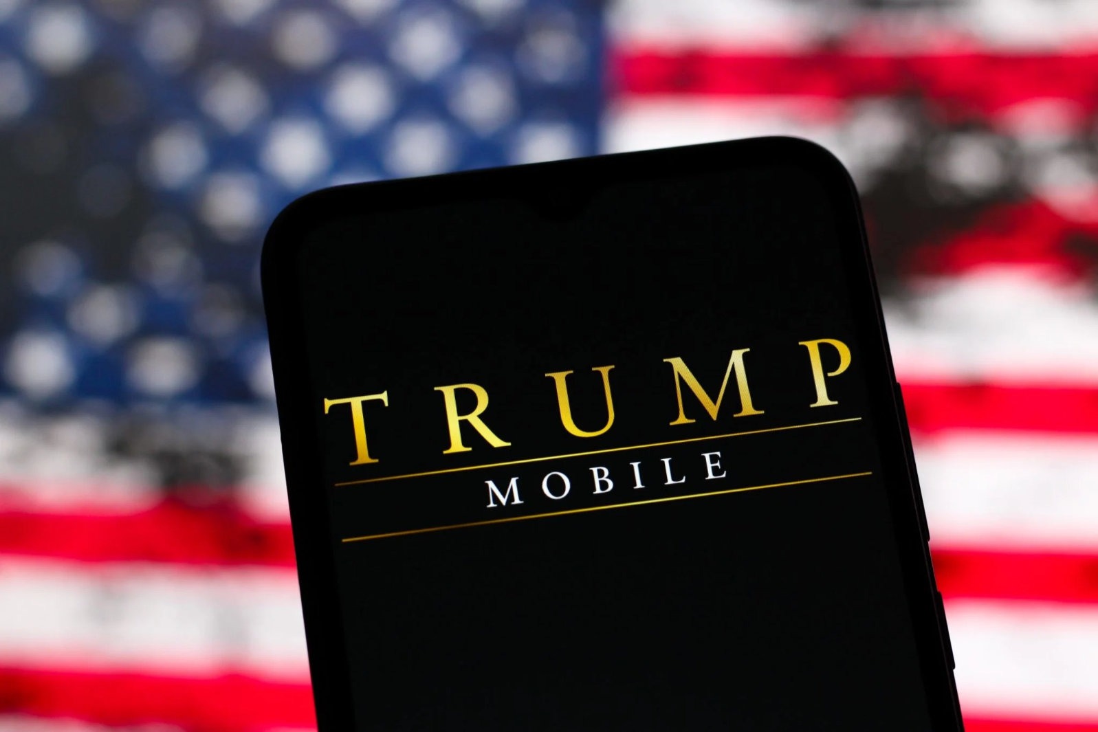 Trump Mobile logo displayed on phone screen with blurry American flag in background