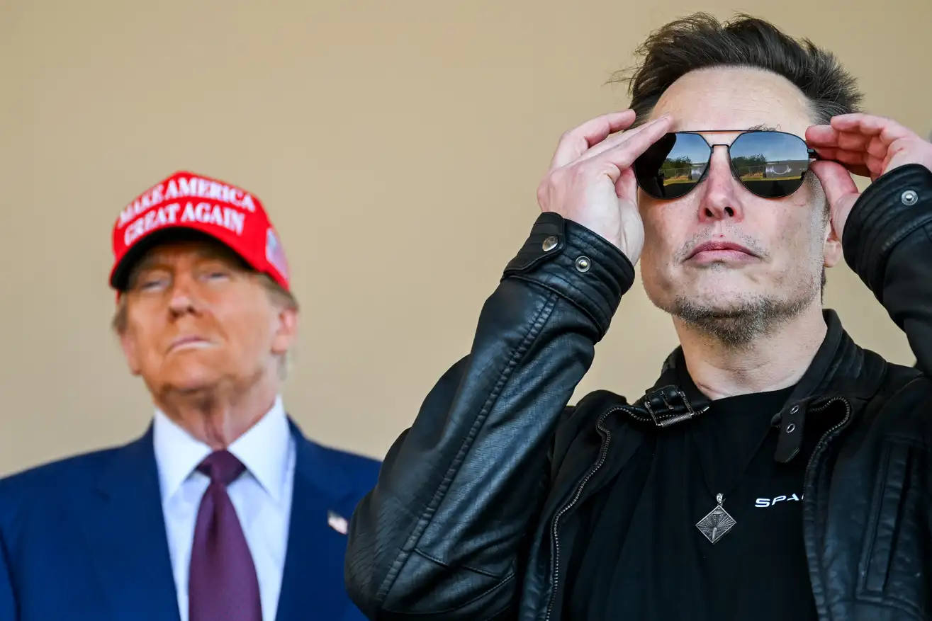 Donald Trump in a red MAGA hat stands behind Elon Musk adjusting his sunglasses, both facing forward with intense expressions.