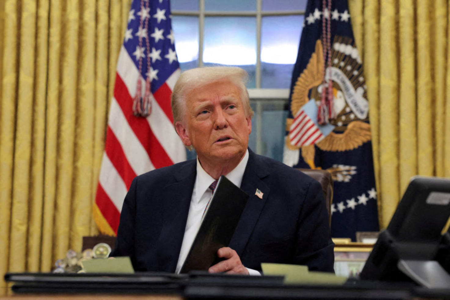 Donald Trump sitting at the Resolute Desk in the Oval Office holding a black folder with flags in the background.