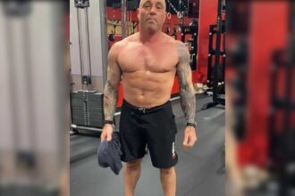 Joe Rogan standing shirtless in a gym holding a t-shirt