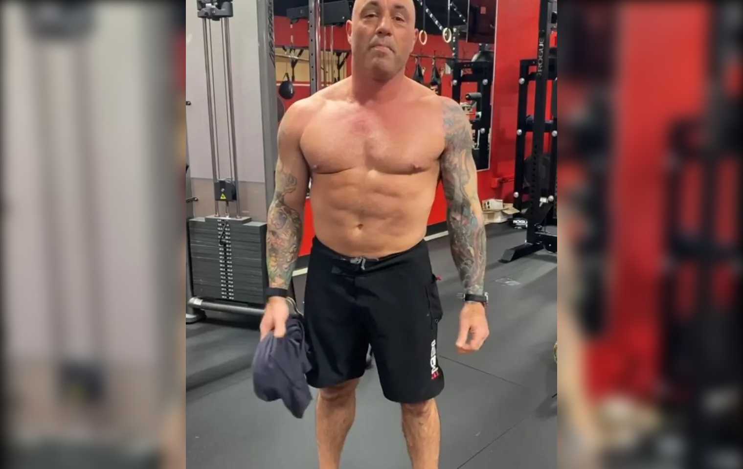 Joe Rogan standing shirtless in a gym holding a t-shirt