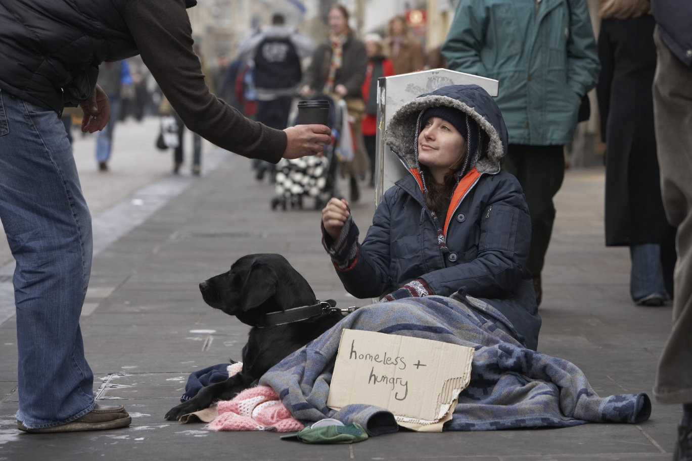 Homeless woman sits on sidewalk with dog while accepting a coffee cup from passerby