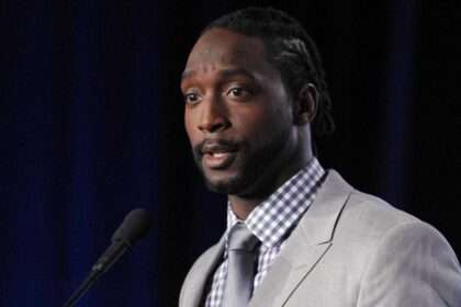 Charles Tillman speaking at a press conference in a gray suit and blue checkered shirt
