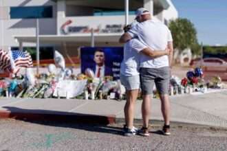 Memorial for Charlie Kirk with flowers, flags, and mourners outside a hospital