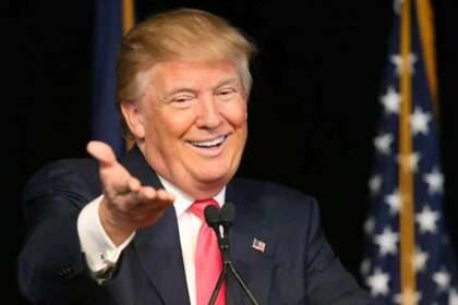 Donald Trump speaking at a podium with a smile and outstretched hand