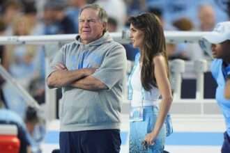 Bill Belichick standing on the sideline at a North Carolina football game beside a woman wearing team colors.