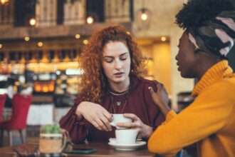 Two women talking over coffee, one appearing apologetic or uncomfortable.