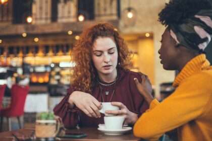Two women talking over coffee, one appearing apologetic or uncomfortable.