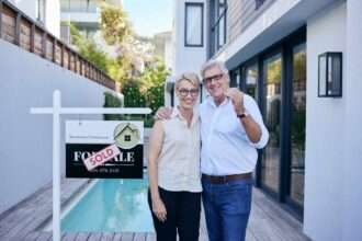 Older couple holding house keys in front of sold sign outside modern home