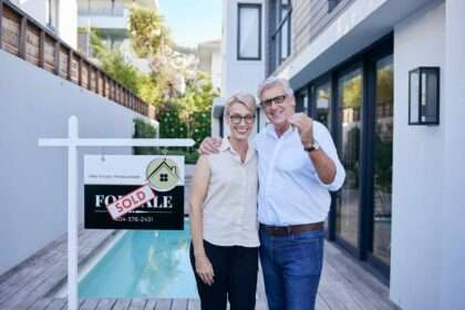 Older couple holding house keys in front of sold sign outside modern home