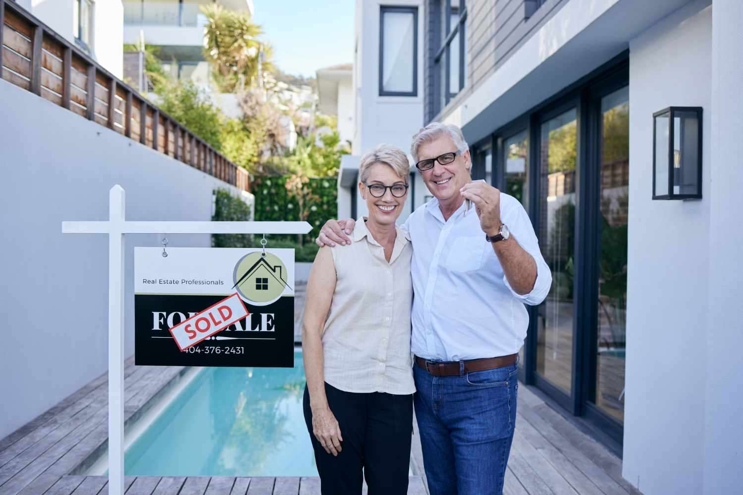 Older couple holding house keys in front of sold sign outside modern home