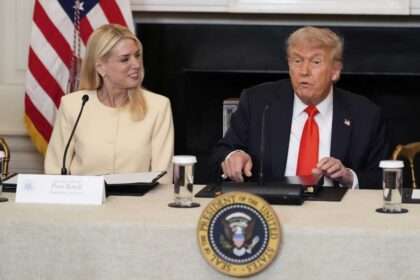 Pam Bondi and President Donald Trump seated at a meeting table with microphones and documents.