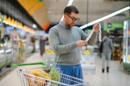 Man reading a long grocery receipt while standing beside a shopping cart filled with food items.