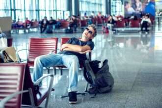 A man sleeping in an airport terminal chair with a backpack beside him.