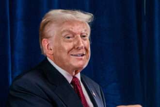 President Donald Trump in a dark suit and red tie, smiling and pointing to his right in front of a blue curtain.