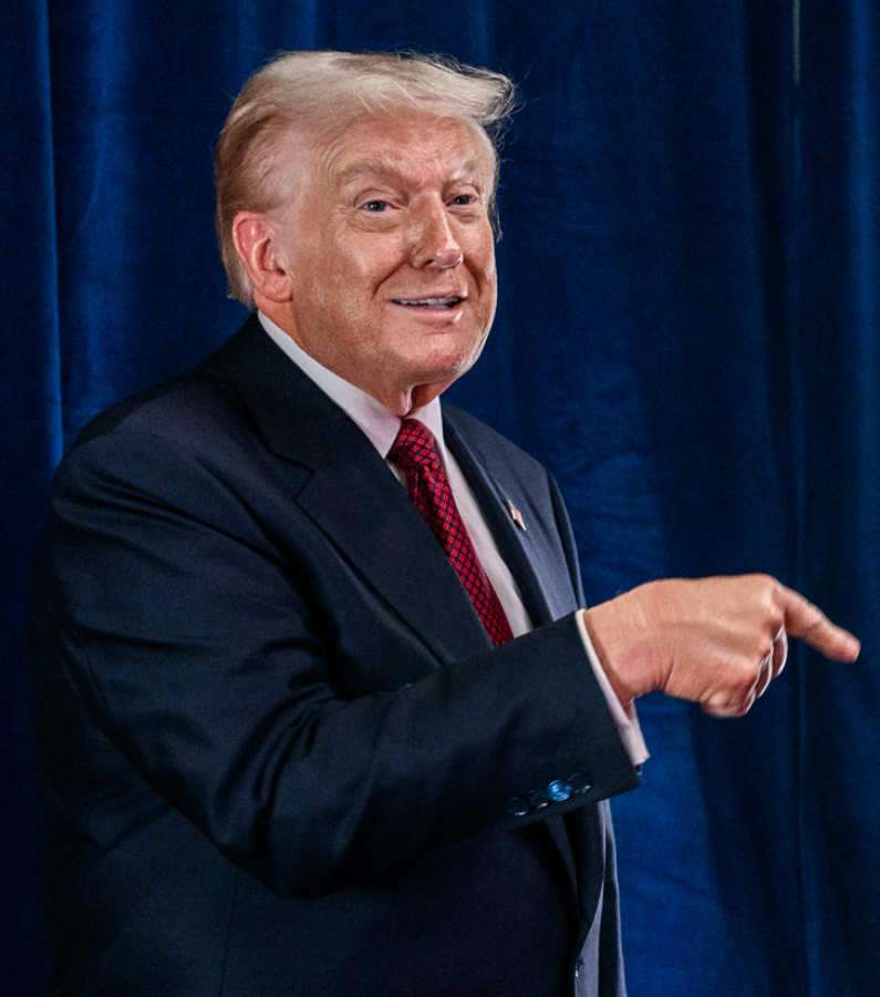 President Donald Trump in a dark suit and red tie, smiling and pointing to his right in front of a blue curtain.