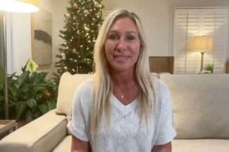 Marjorie Taylor Greene sitting on a beige couch in a living room with a lit Christmas tree behind her, speaking to the camera.