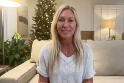 Marjorie Taylor Greene sitting on a beige couch in a living room with a lit Christmas tree behind her, speaking to the camera.