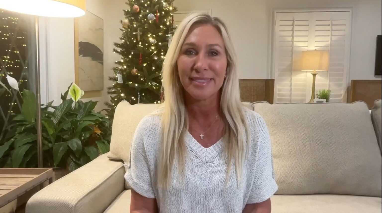 Marjorie Taylor Greene sitting on a beige couch in a living room with a lit Christmas tree behind her, speaking to the camera.