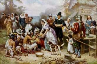Historical painting showing Pilgrims and Native Americans sharing food at an outdoor feast during the first Thanksgiving.