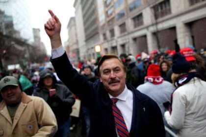 Mike Lindell raising his arm during a public rally in a city street.